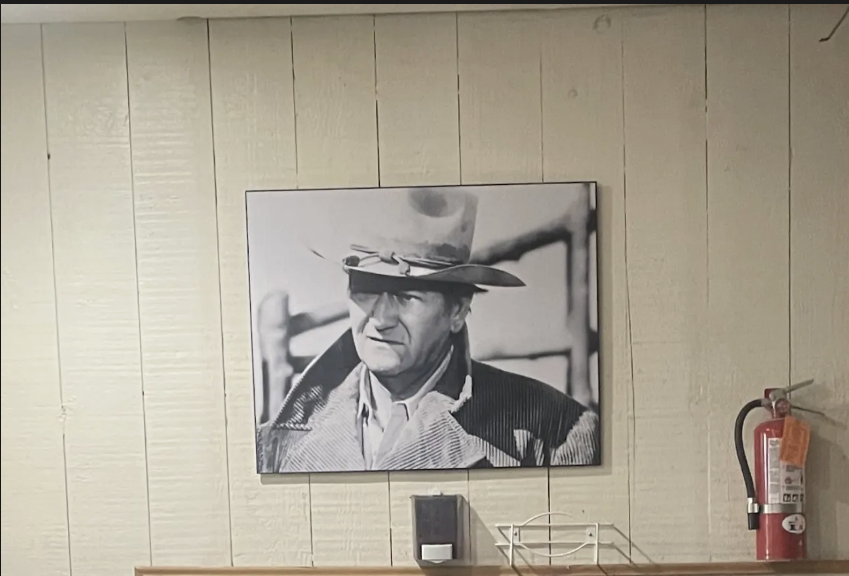 Photo of John Wayne hanging on a wall behind the bar of the Coto Valley Country Club located in California. It symbolizes his prominence, but also his loss of public respect as he's hanging above a pile of discarded boxes.